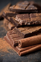 Chocolate bar pieces heap witn cinnamon powder and sticks. Broken chocolate stacked on black background.