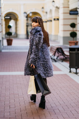 gorgeus smiling brunette with the long straight hair wearing in a fur coat and the white skirt standing against the background of  the classic style building with the shopping bags