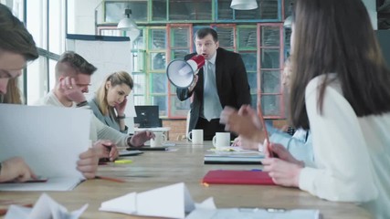 The manager in a business suit gets up from a chair, takes a megaphone in his hand, swears and shouts at the staff. The business team has a bad mood. Office life. Creative interior. Coworking - Powered by Adobe