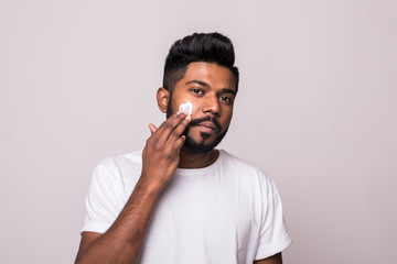 Portrait of satisfied young indian man applying facial cream isolated over white background