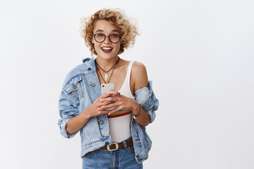 Girl turn attention at camera, distracted from smartphone smiling friendly and happily holding mobile phone as, taking selfie in mirror, showing friends new outfit, standing in dressing room of mall