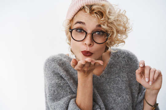 Close-up shot of cute beautiful blond curly-haired woman in glasses beanie and winter sweater sending warm kiss and congratulate with christmas holidays holding palm near folded lips giving mwah