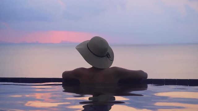 Woman relaxing at the edge of infinity swimming pool. Young Female in Hat watching beautiful Sunset View. She is enjoying vacation at resort by sea. Pink and violet sunset with water reflection 4K