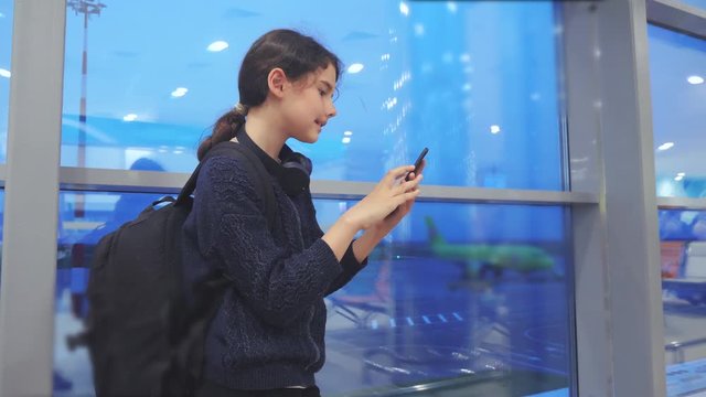 daughter traveler with a backpack airplane flights airport concept. girl teenager with a smartphone looks out the window at the airport area airplanes and bus at a distance. teen girl looks at the