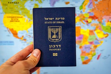 Hand holding the passport of the State of Israel against the colorful world map atlas. Israel citizenship concept, Israeli biometric "darkon" passport illustrative image. Global travel concept.