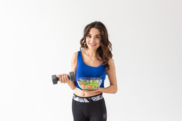 Naklejka premium Healthy lifestyle, diet, fitness and people concept - young woman is eating a salad and doing sport with dumbbell on white background
