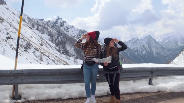 Two young women tourist traveling. Standing on the road and looking around holding a map in Dolomites with big backpack and guitar.