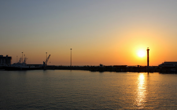 Sunset on sea. The sun painted the sky and water red and yellow. In the distance the dark line of the pier dock. It shows the silhouettes of boats, vessels, crane. Summer is warm, the horizon.