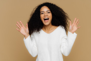 Emotional glad African American woman with crisp black hair, raises hands, gestures actively, observes something wonderful and awesome, wears soft white jumper, models over brown background.