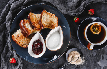 Breakfast with sweet homemade buns on plate over dark background, yogurt, strawberry jam, a cup of tea. Bakery, pastry, dessert, top view.