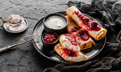 Crepes with cottage cheese, fruit jam, cherries, sour cream and icing sugar on black plate over...