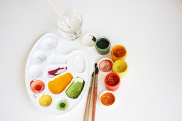 On a white background are art supplies: gouache paints of yellow, red and green colors, a plastic palette, brushes of squirrel fur and a glass with water for washing brushes.