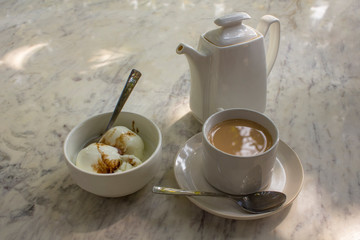 two ice cream scoops and a cup with a milk drink on a saucer with a spoon near a white ceramic teapot on a marble table