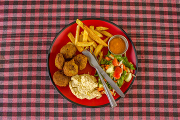 falafel, potatoes, hummus, salad, sauce and fork with a knife in a red plate on a checkered red white black tablecloth top view