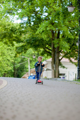 Little boy riding scooter in city park in aummer. Kids sports outdoors. Happy child playing with his scooter. Kid learn to ride scooter in park. Happy childhood