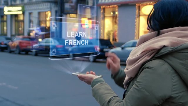 Unrecognizable woman standing on the street interacts HUD hologram with text Learn French. Girl in warm clothes uses technology of the future mobile screen on background of night city