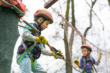 Two cute happy young children, boy and girl in protective harness, carbine and safety helmets on rope way on bright sunny day on green foliage bokeh background. Outdoors activities, games concept.