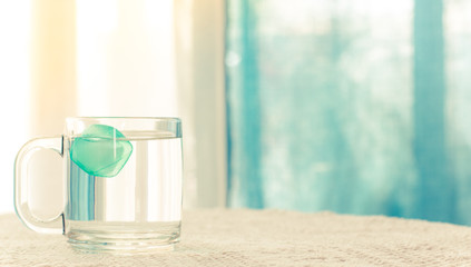 cold fresh drink in hot summer time picture, cup of water with blue peace of ice on a table in white pastel interior environment curtains background, wallpaper pattern copy space