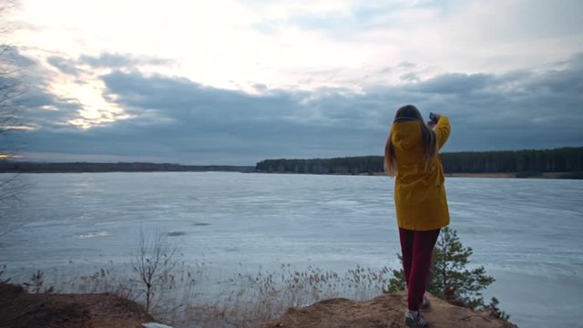 Woman with long hair in a yellow jacket goes to the cliff and takes pictures on her smartphone a beautiful blue sky with clouds and a forest on the other side of a frozen lake. Spring trip, nature.