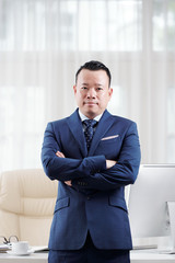 Portrait of Asian confident mature businessman standing in suit with his arms crossed and looking at camera near his workplace at office