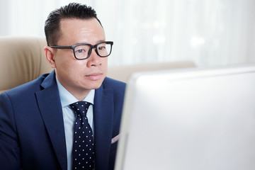 Serious Asian businessman in eyeglasses sitting in front of computer and looking at computer...