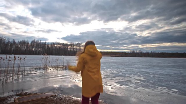 On the shore of a frozen lake, a woman with long hair in a yellow jacket takes pictures of the overcast sky with clouds, and jumps, rejoicing in a successful photo snapshot. Spring trip.