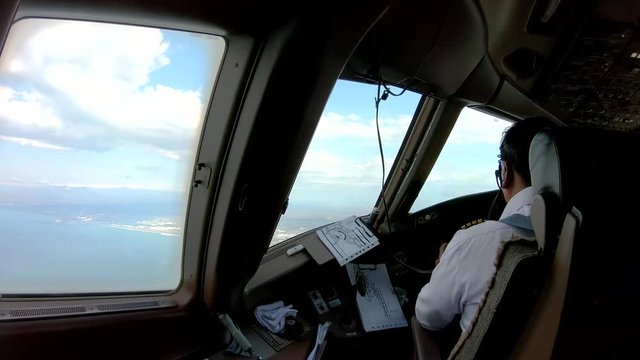 Airline captain controlling the airplane flying over the sea to land on runway in front of him. Outside cockpit is beautiful view of landscape. Seen from inside cockpit behind pilot seat.