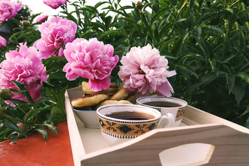 Tea in country style in summer garden in the village. Two cups of black tea on wooden tray and blooming peony flowers in sunlight.