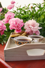 Tea in country style in summer garden in the village. Two cups of black tea on wooden tray and blooming peony flowers in sunlight.