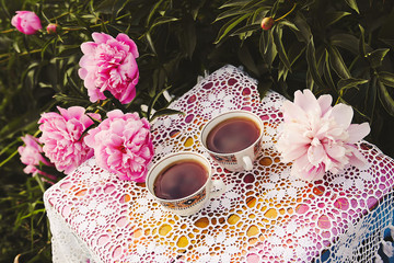 Obraz na płótnie Canvas Tea in country style in summer garden. Two cups of black tea on handmade crocheted vintage lacy tablecloth and blooming peony flowers in sunlight.