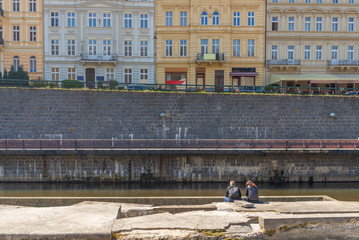Outdoor sunny view, couple or friend sit on the concrete riverbank and background of embankment, street and range of typical European building in Karlovy Vary, Czech Republic. Chilling and relax mood.