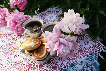 Tea in country style in summer garden. Two cups of black tea and pancakes on handmade crocheted vintage lacy tablecloth and blooming peony flowers in sunlight.
