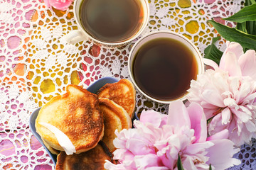 Tea in country style in summer garden. Two cups of black tea and pancakes on handmade crocheted vintage lacy tablecloth and blooming peony flowers in sunlight.