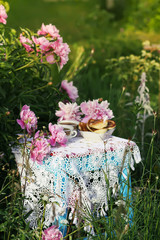 Tea in country style in summer garden. Two cups of black tea and pancakes on handmade crocheted vintage lacy tablecloth and blooming peony flowers in sunlight.
