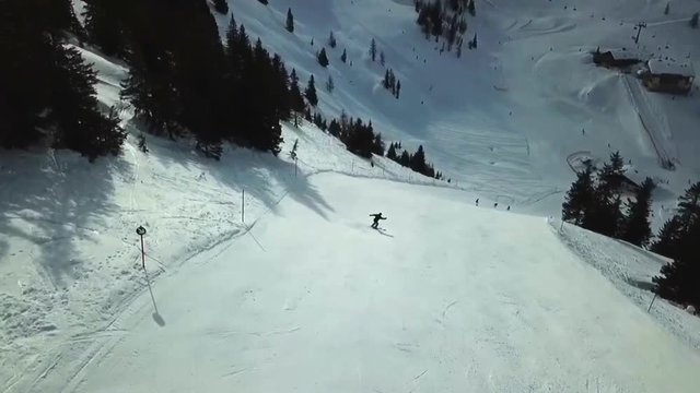 Drone shot above a ski slope in Flachau, Austria, with people skiing down the snowy mountains near some trees, on a sunny day