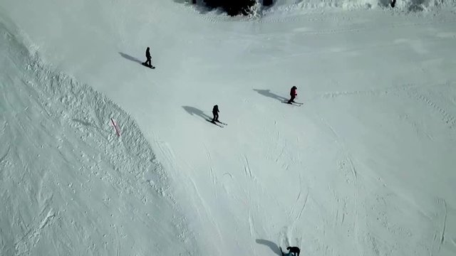 Drone shot above a ski slope in Flachau, Austria, with people skiing down the snowy mountains near some trees, on a sunny day