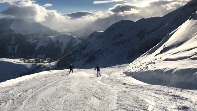 Point of view shot skiing down a ski slope in Flachau, Austria, behind other people, with snowy mountains and some trees in the background, on a sunny evening with some clouds