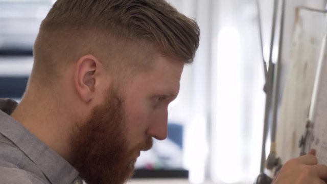 Closeup full bearded Caucasian civil engineer drawing blueprints. Close-up horizontal Steadicam tracking shot. Male worker with beard indoors sketching, drawing board.