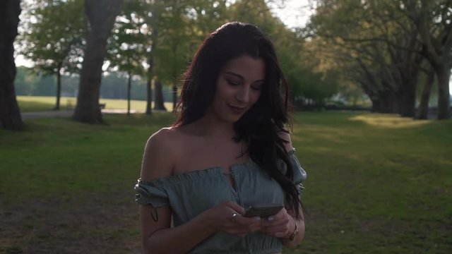 Attractive young brunette latina woman in a casual outfit, uses her smartphone, smiles happily to the text messages, looks around.