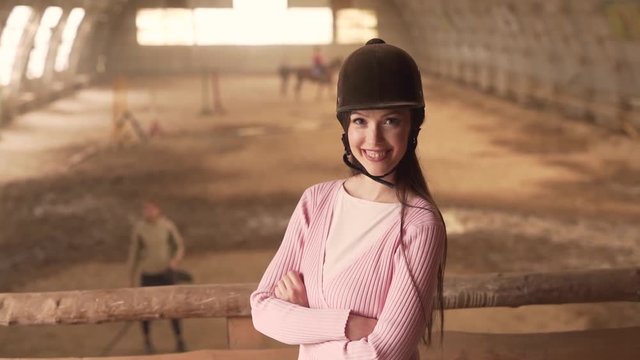 Young Beautiful Caucasian Female With Equestrian Helmet on Head Standing By Wooden Fence in Horse Hangar Stable Looking And Posing To Camera. Blurred Background