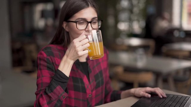Upper body part close up video of beautiful female with glasses in red and black checked shirt. She works on her laptop and stops for seizing moment. She starts drinking cup of tea and enjoys herself