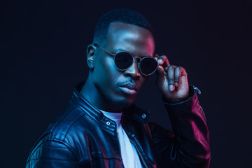 Young African male pictured against dark background in black jacket and trendy sunglasses