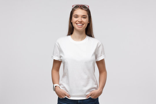 Young smiling woman standing with hands in pockets, wearing blank white t-shirt with copy space for your logo or text, isolated on gray background