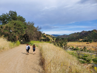 Woman and daughter walking together on a trail or dirt road in the woods next to a yellow field