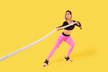 Fototapeta na wymiar Strong woman pulling rope. Photo of sporty attractive woman in pink and black sportswear isolated on yellow background. Strength and motivation.