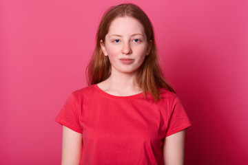 Fototapeta premium Half lengh portrait of young student female looks with calm facial expression at camera, wearing red casual t shirt, has long straight brown hair and beautiful blue eyes, isolated over pink background