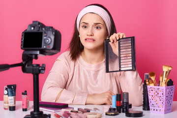 Indoor shot of disappointed attractive beaty blogger, holding palette of eyeshadow, demonstration of it to viewers, posing isolated over pink background in studio in front of camera, looks upset.