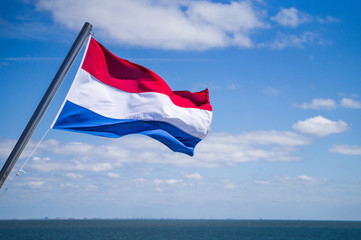 Waving flag of the Netherlands fixed to yacht mast with blue sky with white clouds and sea on background 