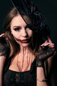Portrait of a sexually beautiful girl in a scary zombie image with a bloody mouth. Halloween makeup, horror party costume. Black witch hat, lace gloves, red smudges on a woman's face.