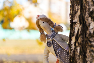 A female Tilda doll in a colorful dress looks out from behind a tree. Registration of holidays. Interior fairy doll handmade. Art and creativity. Blurred background